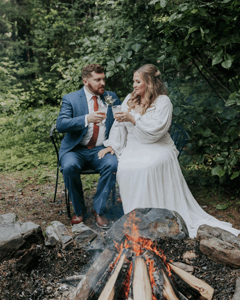 groom and bride sitting by campfire sipping wine at cabin in seward alaska elopement